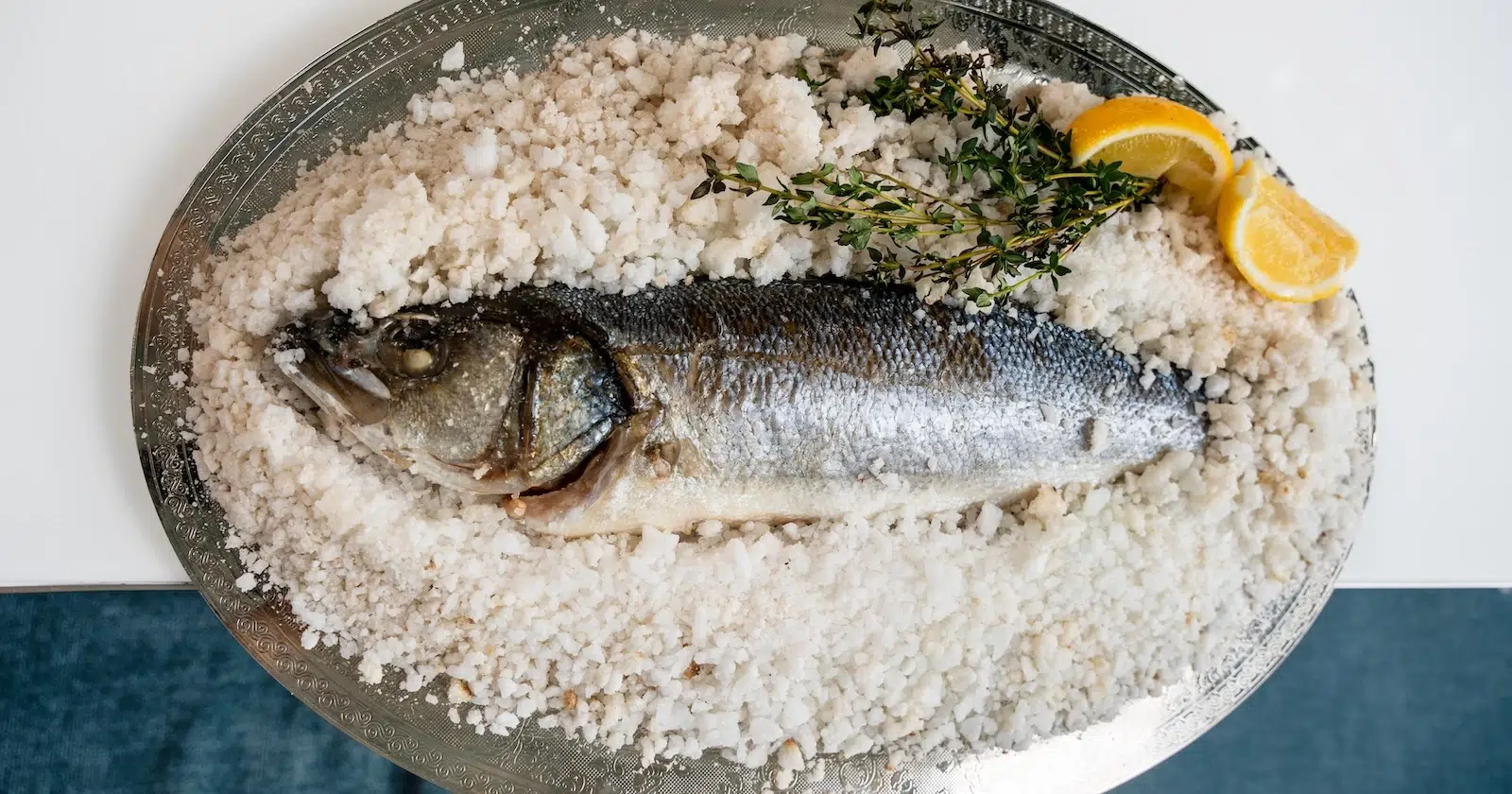 A beautifully baked whole fish encased in a salt crust is presented on a silver platter. The dish is garnished with fresh thyme and lemon wedges, adding a vibrant and aromatic touch.