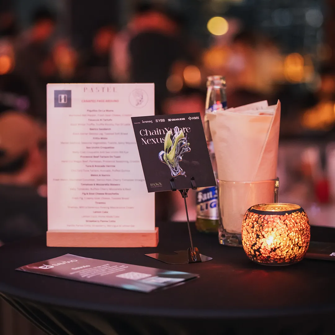 A close-up of a table at Pastel Bangkok with a menu, a candle, a glass with napkins, and a small event card, set against a blurred restaurant background.